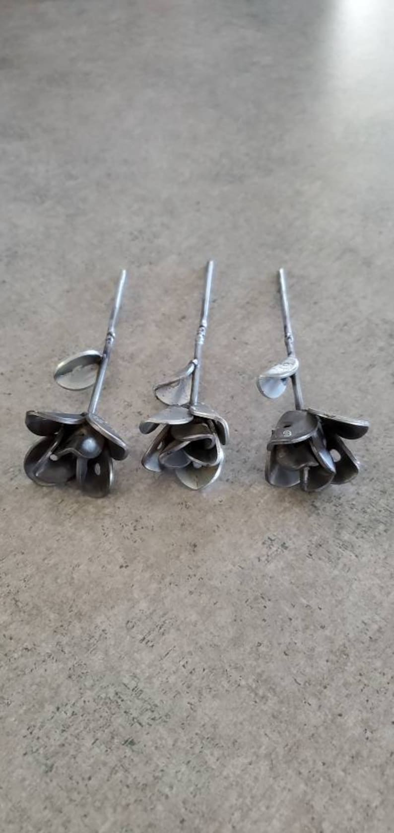 May include: Three metal roses with stems, each crafted from recycled metal, are arranged on a gray surface. The roses are a unique and artistic way to add a touch of nature to any home.