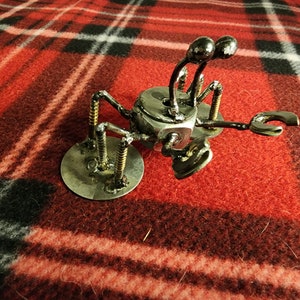 May include: A metal sculpture of a crab with a rounded body and multiple legs made from screws and nuts. The crab is sitting on a red and black plaid surface.