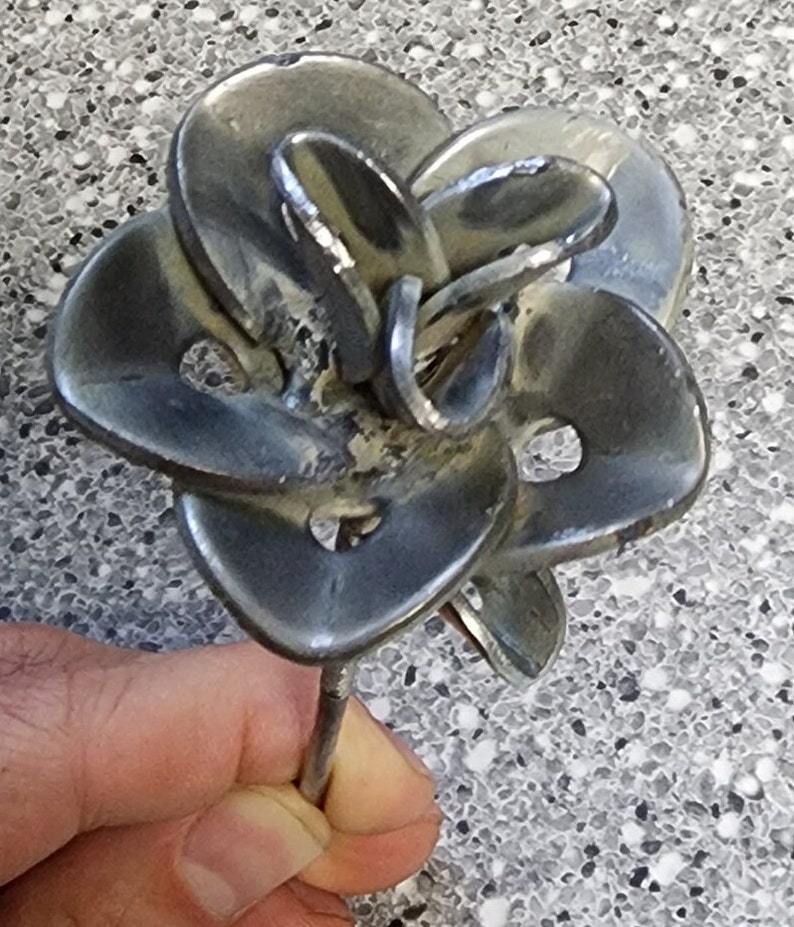 May include: A metal sculpture of a flower with multiple petals, crafted from recycled metal. The flower is silver in color and has a long stem.