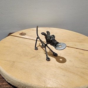 May include: A metal sculpture of a dog made from nuts, bolts, and washers. The dog is standing on a wooden surface and has a washer in its mouth.