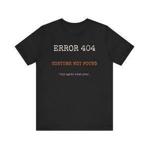 Op de afbeelding: Een zwart t-shirt met korte mouwen voorzien van witte tekst ERROR 404, oranje tekst COSTUME NOT FOUND en kleinere witte tekst eronder die > try again next year_ leest in een typemachine-stijl lettertype gecentreerd op de borst.