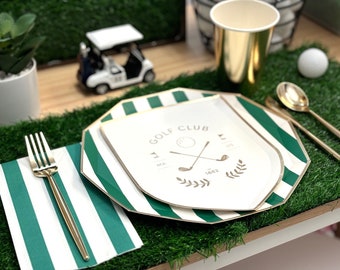 May include: A golf-themed party setting with green and white striped plates, napkins, and a gold fork and spoons. A paper plate with "Golf Club" text and golf club graphic is on top. A toy golf cart and golf ball are also present.