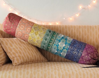 May include: A colorful, patterned yoga bolster with a strap, resting on a beige couch with a pillow. A small green plant is in a pot on a table in the foreground.