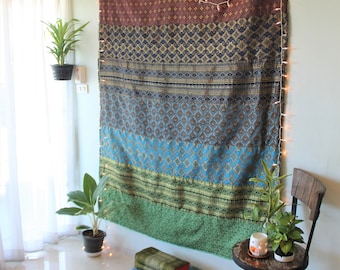 May include: A large rectangular wall hanging featuring horizontal bands of intricate woven patterns in shades of brown, blue, and green. The fabric is draped against a wall and decorated with a string of small white lights along the edges.