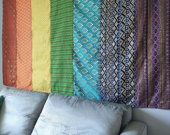 May include: A colorful tapestry with a rainbow of colors and geometric patterns. The tapestry is hanging on a wall above a gray couch.