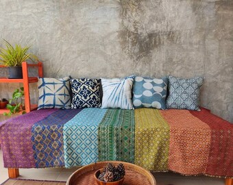 May include: A colorful, patterned throw blanket with a rainbow of colors, including purple, blue, green, yellow, orange, and red, is draped over a wooden bench. The bench has six pillows with various blue and white patterns. The bench is in front of a concrete wall.
