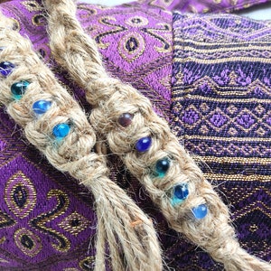May include: Two braided jute cords with colourful glass beads woven into the braid. The cords are on a purple and gold fabric background.