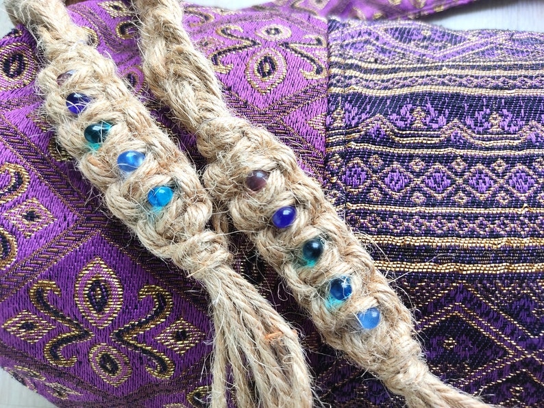 May include: Two braided jute cords with colourful glass beads woven into the braid. The cords are on a purple and gold fabric background.