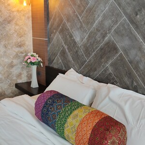 May include: A colorful cylindrical pillow with a rainbow pattern. The pillow is made of fabric with a variety of textures and designs. It is lying on a white bed with white pillows.