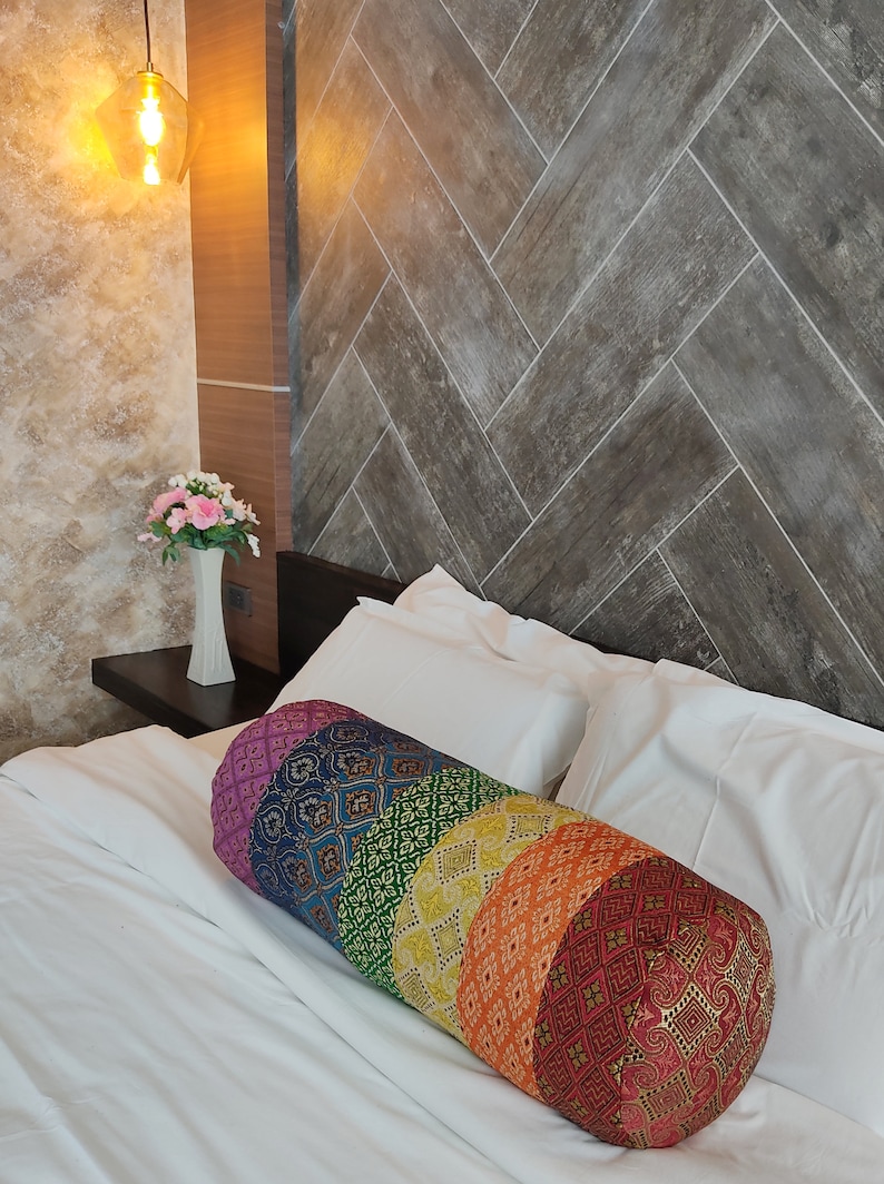 May include: A colorful cylindrical pillow with a rainbow pattern. The pillow is made of fabric with a variety of textures and designs. It is lying on a white bed with white pillows.