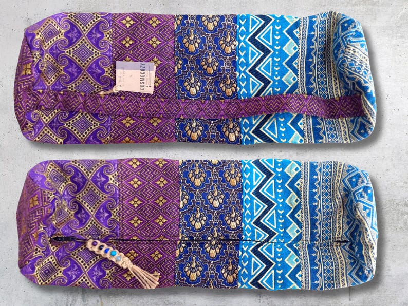 May include: Two purple and blue patterned yoga bolsters with a gold trim. The top bolster has a tag that reads "Cosmic Cozy".