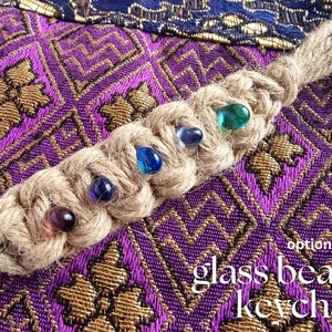 May include: A braided jute keychain with a metal ring and colorful glass beads. The keychain is on a purple and gold patterned fabric background. The text "glass beads keychain" is written in white on the fabric.