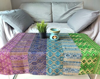 May include: A colorful table runner with a geometric pattern in shades of purple, blue, and green. A teal ceramic oil burner with a candle sits on the table runner.
