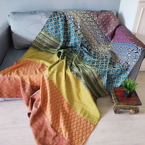 May include: A colourful patchwork throw blanket with a geometric pattern in shades of blue, green, yellow, orange, and red. The blanket is draped over a grey sofa and a small wooden table with a potted plant.