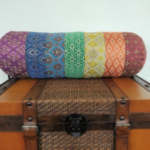 May include: A colorful cylindrical pillow with a rainbow pattern of purple, blue, green, yellow, and orange fabric. The pillow is sitting on top of a wooden chest with a woven lid.