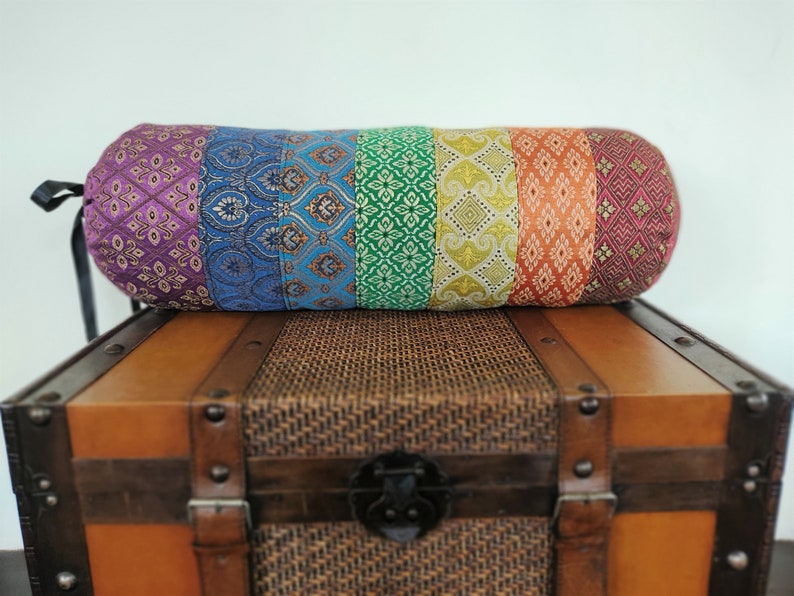 May include: A colorful cylindrical pillow with a rainbow pattern of purple, blue, green, yellow, and orange fabric. The pillow is sitting on top of a wooden chest with a woven lid.