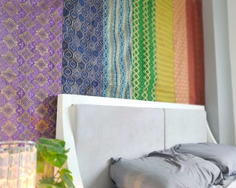 May include: A bedroom scene featuring a white headboard and gray bedding. Behind the headboard, colorful fabric panels in shades of purple, blue, green, yellow, orange, and red create a vibrant backdrop. The fabrics have intricate gold patterns.