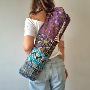May include: A colourful yoga mat bag with a patchwork design featuring purple, blue and gold patterns. The bag has a long strap and a tassel.