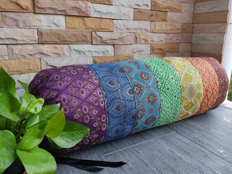 May include: A colorful cylindrical pillow with a rainbow pattern. The pillow is made of fabric with a variety of textures and patterns. The pillow is lying on a wooden surface.