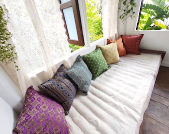 May include: A white linen daybed with a variety of patterned throw pillows. The pillows are in shades of green, yellow, purple, and orange. The daybed is situated near a window with white lace curtains.