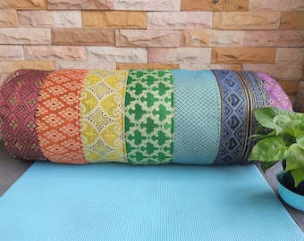 May include: A colorful cylindrical pillow with a rainbow pattern. The pillow is made of fabric with different geometric designs in shades of red, orange, yellow, green, blue, and purple. The pillow is resting on a blue yoga mat.
