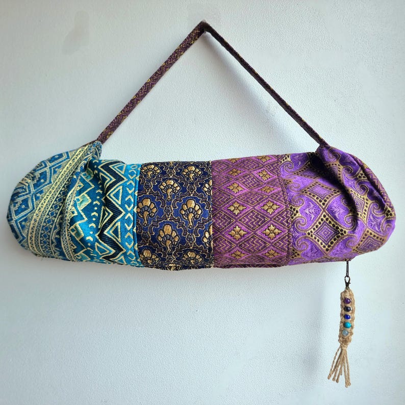 May include: A multi-colored yoga mat bag with a purple, blue, and gold patterned design. The bag has a brown strap and a tassel with beads.