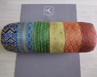 May include: A multi-colored cylindrical yoga bolster with a patterned fabric design. The bolster is made of six different fabric panels, each with a unique geometric pattern. The panels are arranged in a rainbow color scheme, starting with a dark blue panel and ending with a red panel.