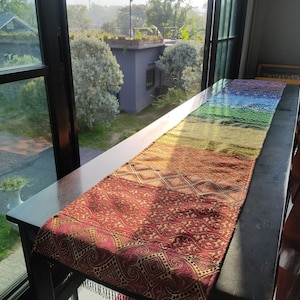 Puede incluir: Un camino de mesa colorido con un diseño de gradiente de arcoíris. El camino de mesa pasa del rojo al naranja, amarillo, verde y azul. La parte inferior presenta un diseño detallado y estampado en tonos rojos y dorados. El camino de mesa se muestra sobre una mesa oscura.