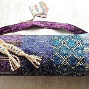 以下が含まれることがあります： 紫、青、金色のヨガマットバッグ。編まれたジュートのストラップとタッセルが付いています。バッグは幾何学模様です。