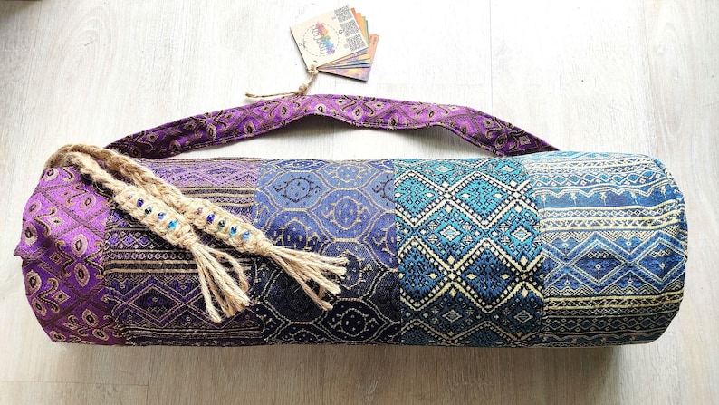 May include: A purple, blue and gold yoga mat bag with a braided jute strap and tassels. The bag has a geometric pattern.