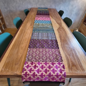 May include: A long rectangular table runner featuring a patchwork design of various woven fabric sections. The patterns include floral, geometric, and striped motifs in vibrant shades of magenta, purple, teal, blue, and gold, finished with black fringe.