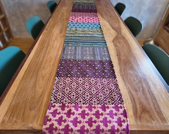 May include: A long rectangular table runner featuring a patchwork design of various woven fabric sections. The patterns include floral, geometric, and striped motifs in vibrant shades of magenta, purple, teal, blue, and gold, finished with black fringe.
