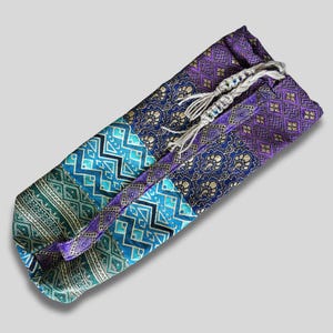 May include: A rectangular fabric pouch featuring a patchwork design of purple, blue, and green panels with intricate gold-toned geometric and floral patterns. The pouch is secured with a braided light-colored cord featuring small decorative beads.