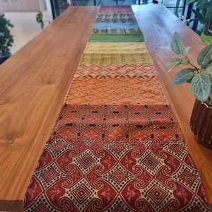 Könnte beinhalten: Ein farbenfroher Tischläufer mit geometrischem Muster. Der Läufer ist aus gewebtem Stoff und hat eine Vielzahl von Farben, darunter Rot, Orange, Gelb, Grün, Blau und Lila. Der Läufer liegt auf einem Holztisch.