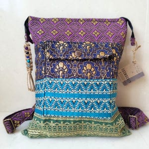 May include: A multi-colored patchwork crossbody bag with a purple, blue, green, and gold pattern. The bag has a flap with a button closure and a small pocket on the front. The bag has a long adjustable strap and a small tassel on the side. The bag has a tag that says "COSMOGROOVY".