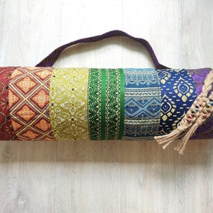 May include: A yoga mat bag in a rainbow of colours. The bag has a variety of patterns and colours, including red, orange, yellow, green, blue, and purple. It has a woven handle and a decorative tassel.