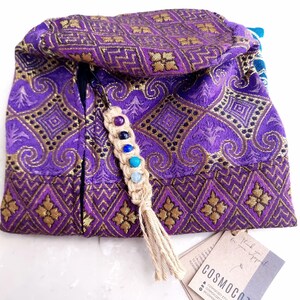 May include: Purple and gold fabric pouch with a zipper closure and a tassel. The pouch has a beaded charm with colorful stones attached to it. The pouch is labeled "COSMOCOZY".