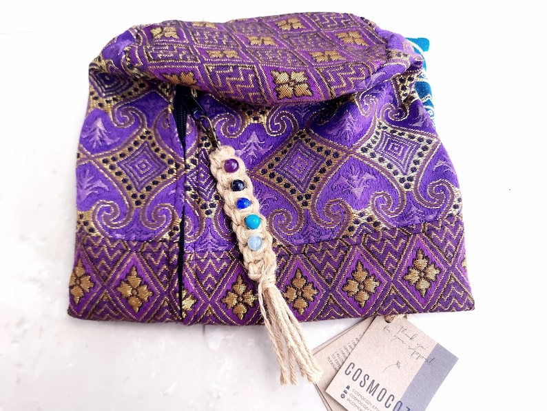 May include: Purple and gold fabric pouch with a zipper closure and a tassel. The pouch has a beaded charm with colorful stones attached to it. The pouch is labeled "COSMOCOZY".
