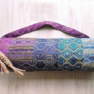 May include: A purple and blue yoga mat bag with a patterned design. The bag has a long strap and a drawstring closure.