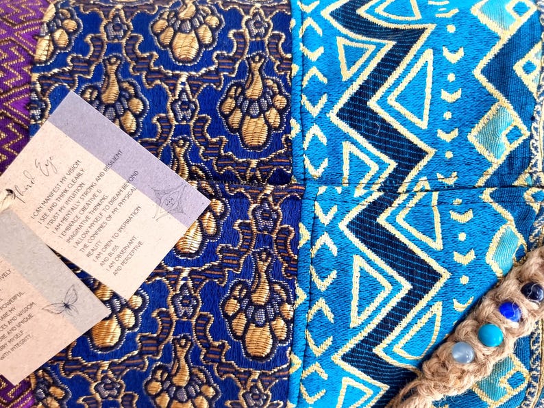 May include: A blue and gold patterned fabric pouch with a brown cord and blue and green stones. The pouch has two tags attached with the text "Third Eye" and "I can manifest my vision, I see and think clearly, I trust my intuition, I am mentally strong and resilient, I embrace creative and imaginative thinking, I allow myself to dream beyond the confines of my physical reality, I am open to inspiration and beauty, I am observant and perceptive."