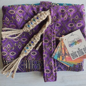 May include: Purple fabric pouch with a gold pattern and a tassel closure. The pouch has a woven cord with colourful beads and a tag that says "COSMOCOZY" with social media icons and QR codes.