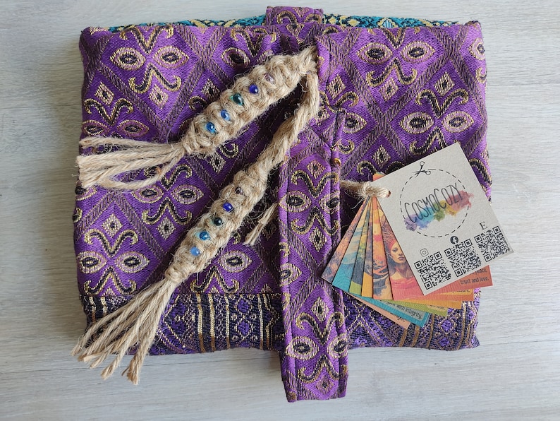 May include: Purple fabric pouch with a gold pattern and a tassel closure. The pouch has a woven cord with colourful beads and a tag that says "COSMOCOZY" with social media icons and QR codes.