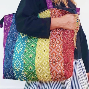 以下が含まれることがあります： 虹色の模様が施された、大きなカラフルなトートバッグ。バッグは布製で、織り込まれたストラップが付いています。バッグは人が持っています。
