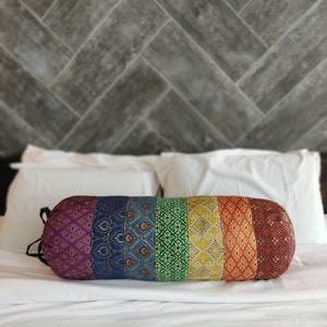 May include: A colorful cylindrical pillow with a rainbow pattern. The pillow is made of fabric with different textures and patterns. It is lying on a white bedspread.