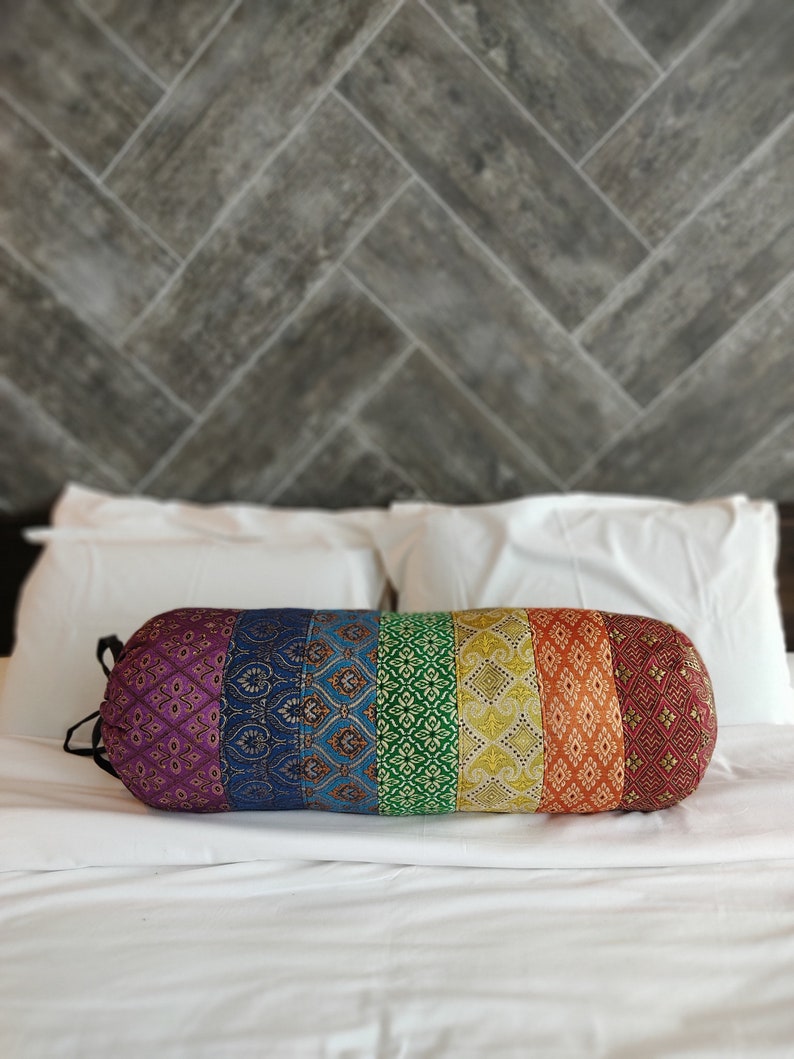 May include: A colorful cylindrical pillow with a rainbow pattern. The pillow is made of fabric with different textures and patterns. It is lying on a white bedspread.