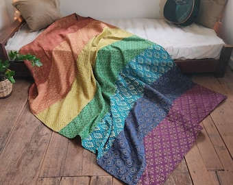 May include: A colorful patchwork quilt with a rainbow of colors, including red, orange, yellow, green, blue, and purple. The quilt is made of different fabrics with intricate patterns and is folded on a wooden floor.