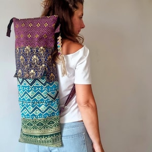 以下が含まれることがあります： 紫、金、青、緑のパターンでデザインされた、マルチカラーのヨガマットバッグ。バッグには長いストラップが付いており、バックパックとして着用されています。