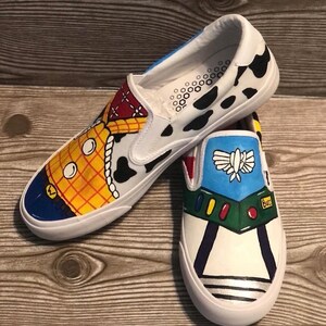 May include: Hand-painted white slip-on shoes with designs from the film Toy Story. One shoe depicts Woody, the other Buzz Lightyear. The shoes have a white sole and a black and white cow print. The shoes are a fun, colourful design.