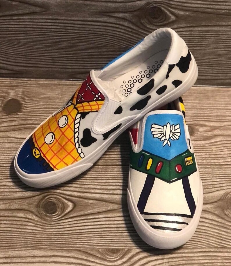 May include: Hand-painted white slip-on shoes with designs from the film Toy Story. One shoe depicts Woody, the other Buzz Lightyear. The shoes have a white sole and a black and white cow print. The shoes are a fun, colourful design.