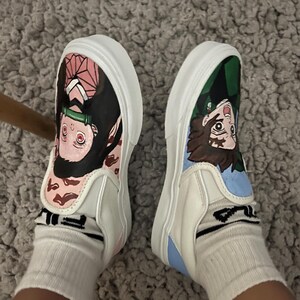 May include: White slip-on trainers, each decorated with anime character artwork. One shoe has pink accents, the other green and blue. The trainers are worn with white socks. The shoes are on a grey carpet.